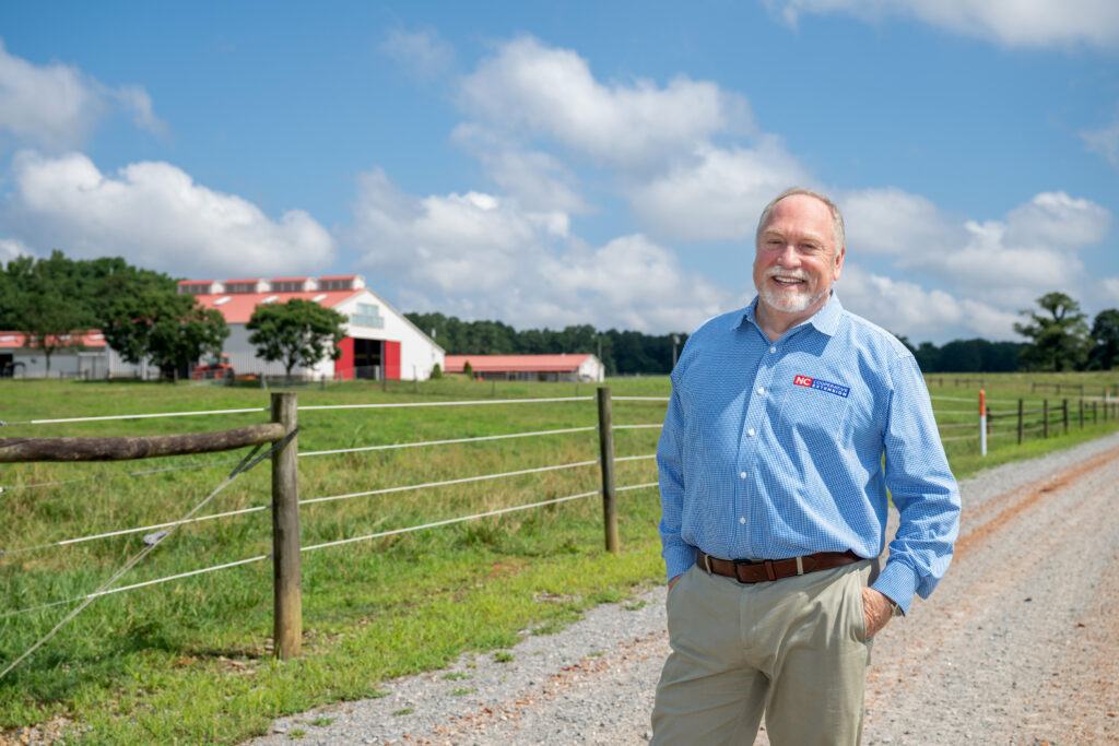 A man in a Cooperative Extension shirt stands on the road to a dairy barn.