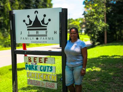 Woman standing by JIREH Family Farms sign reading BEEF, PORK CUTS, CHICKEN, FRESH EGGS