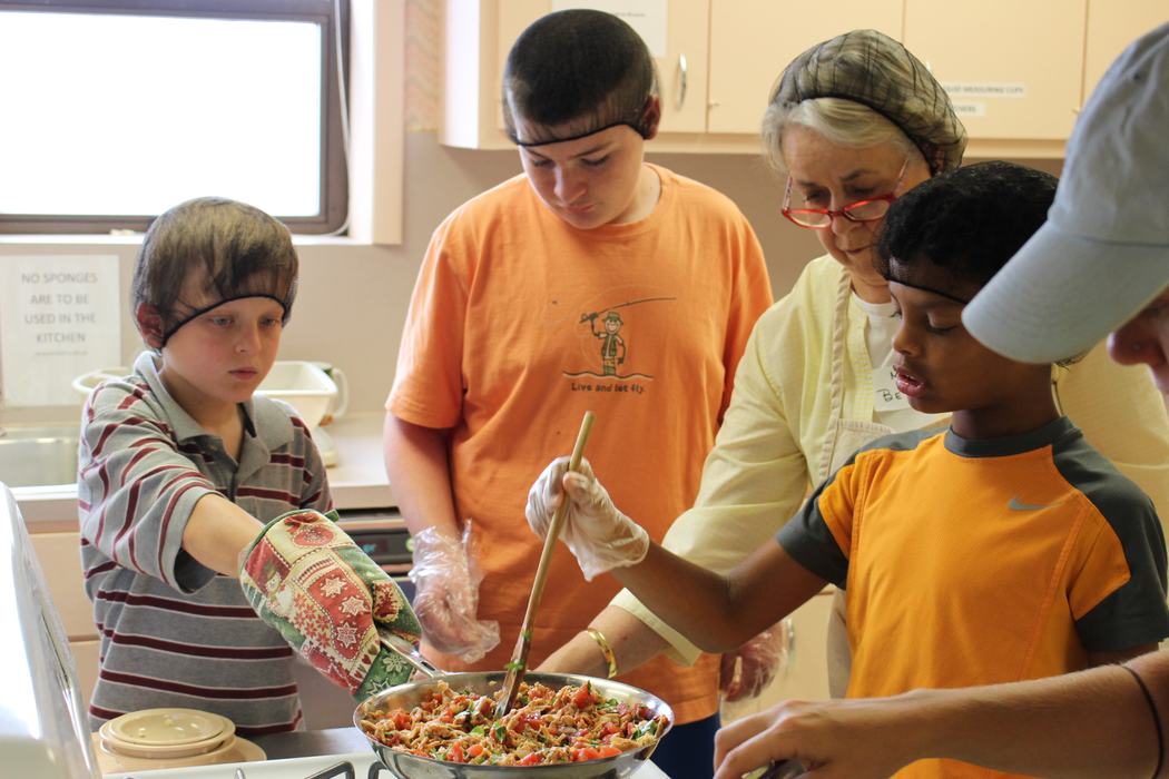 Children and an adult wearing hairnets and gloves work together to cook a colorful vegetable dish in a kitchen setting.