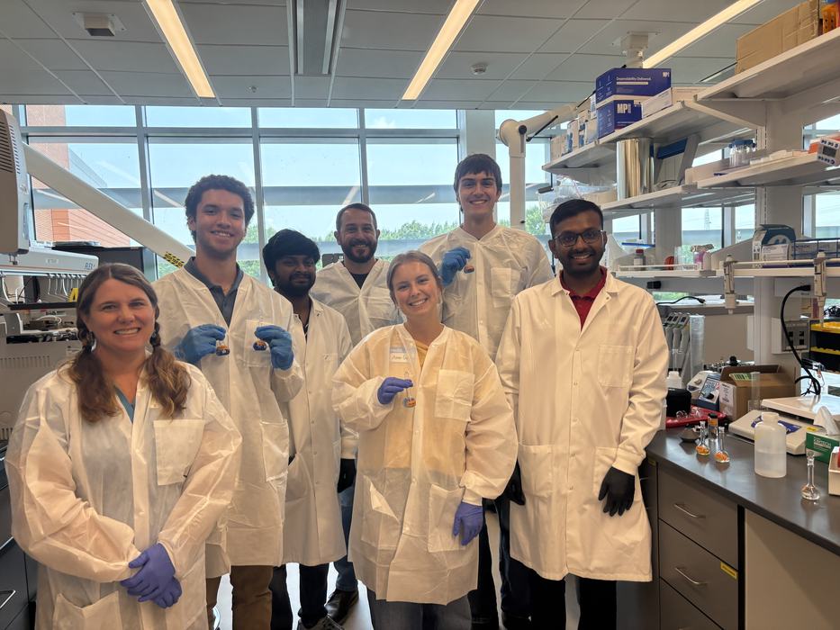 A group of seven people wearing lab coats and gloves smile for a group photo inside a laboratory.
