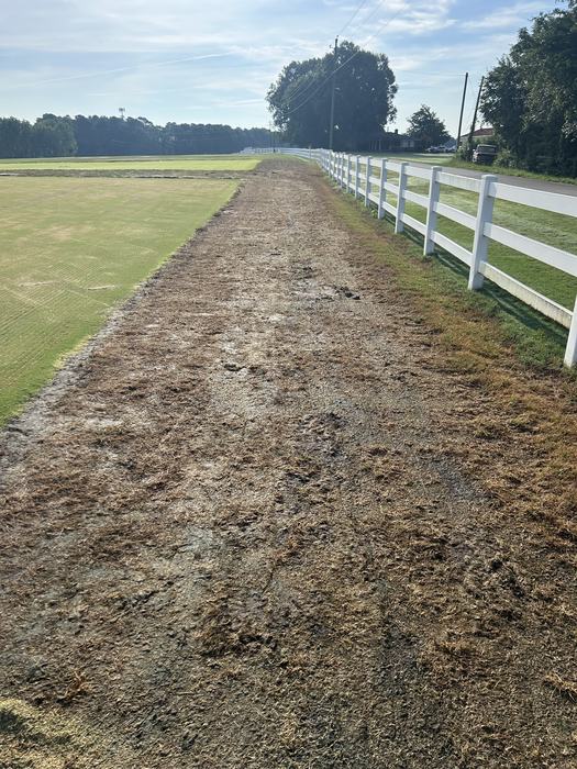 Dirt walking path beside a mowed field with a white fence running right to the horizon