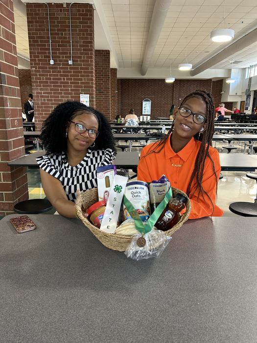Two girls - one wearing an orange shirt and one wearing a black and white striped dress - sitting at a table in a cafeteria. A basket sits in front of them, filled with non-perishables, with a bronze medal and a white ribbon saying "State" in green letters resting on top.