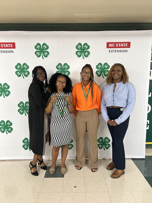 Four girls - two wearing dresses and two wearing dress pants - stand in front of a white background decorated with the official 4-H clover emblem and the Cooperative Extension banner. Three girls are wearing bronze medals. 