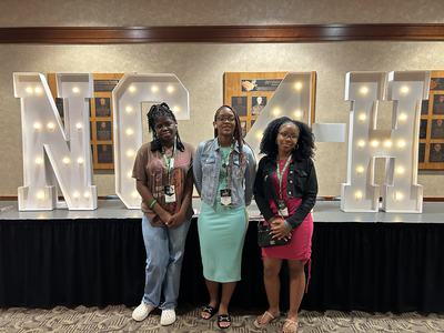 Three girls—two in dresses and one in jeans—standing in front of large 'NC 4-H' letters at NC 4-H Congress.