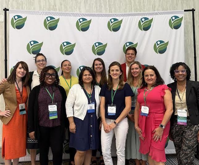 Group photo of thirteen professionals standing in front of a backdrop with a green and blue leaf logo, wearing conference badges and smiling for the camera.