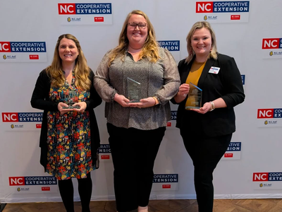 Three women holding awards in front of NC Cooperative Extension backdrop