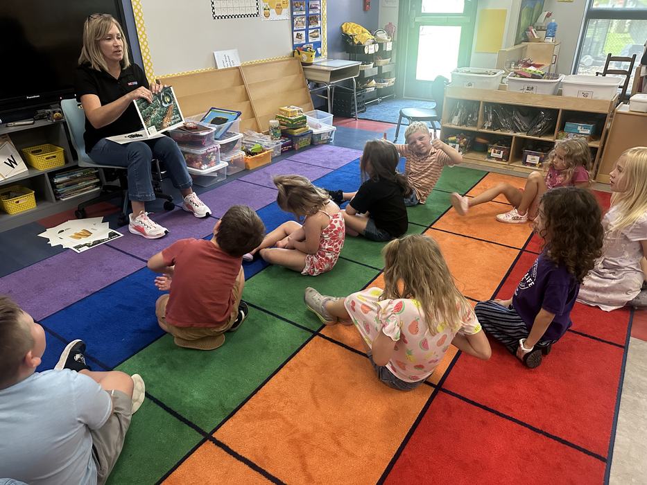 A woman reads a picture book to a group of young children seated on a colorful classroom rug.