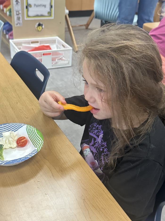 A girl bites into a slice of orange bell pepper while seated at a table with a plate of fresh vegetables.