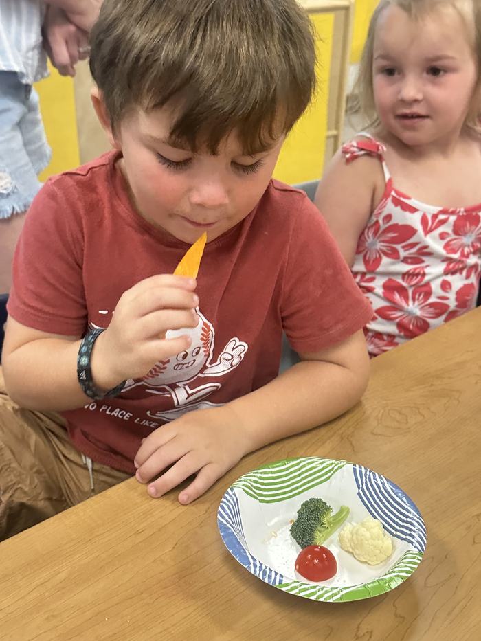 A young boy sits at a table holding a vegetable slice near his mouth, with a plate of raw vegetables in front of him and another child watching.