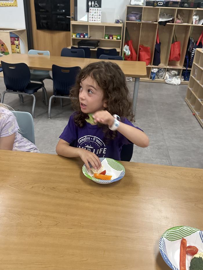 A girl in a purple shirt holds a piece of broccoli while sitting at a classroom table with a paper plate of cut vegetables.