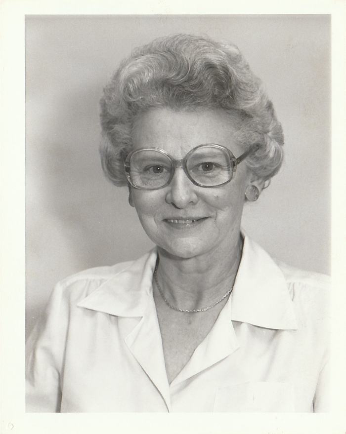 Older woman with short curly hair, large eyeglasses, necklace, and white collared blouse.