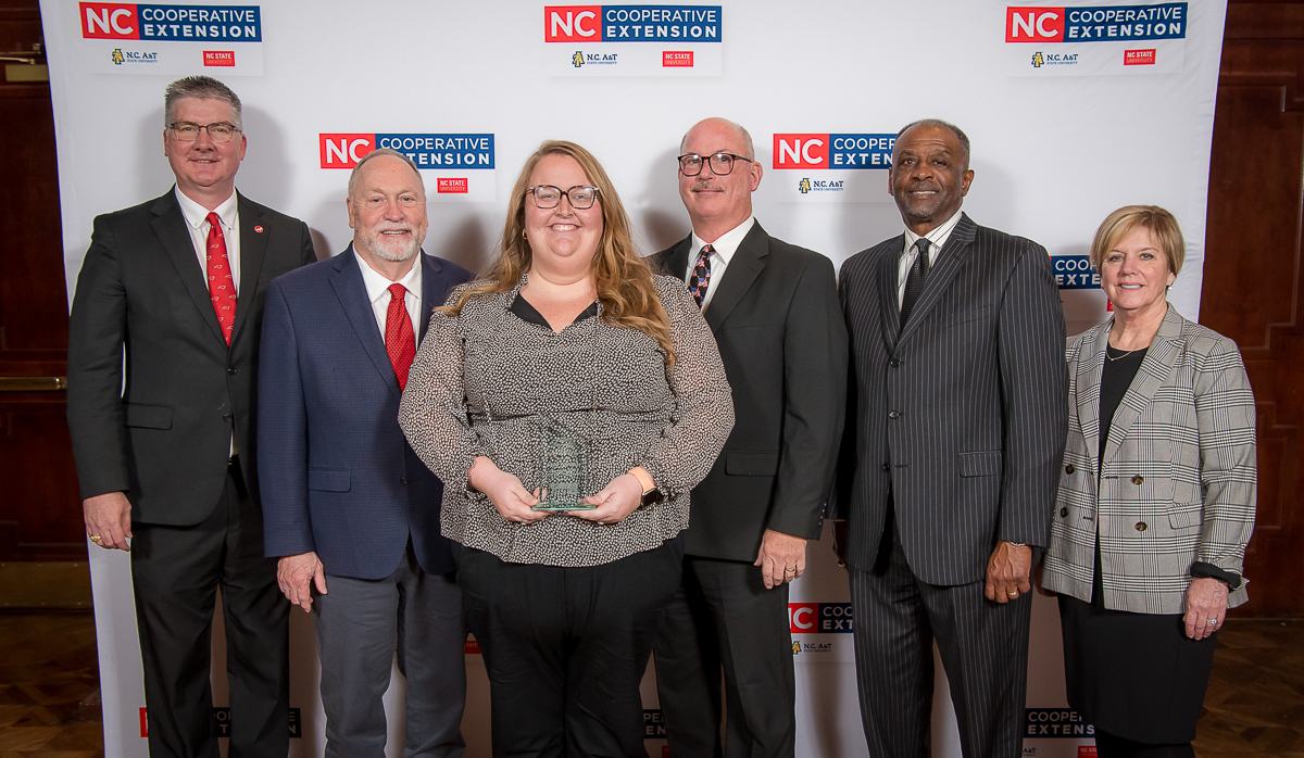 Six people in suits; center woman holds glass award in front of NC Cooperative Extension banner
