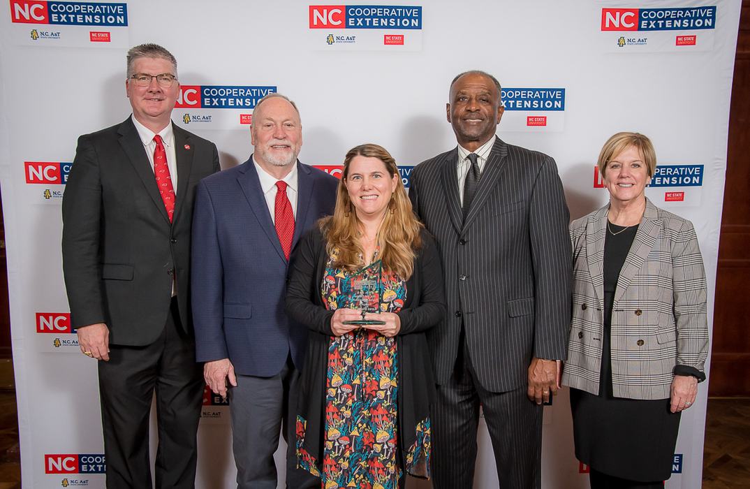 Five people standing in front of NC Cooperative Extension backdrop; center woman holding award
