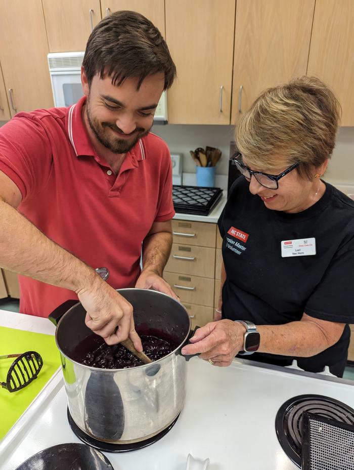 A man and woman stir down a jam in a tall pot.