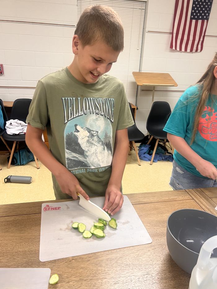 Young person slicing cucumbers on a cutting board; shirt reads "YELLOWSTONE."