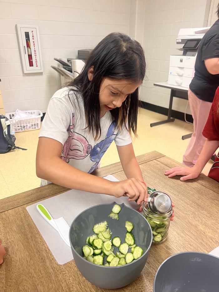 Girl placing sliced cucumbers from a bowl into a glass jar on a table