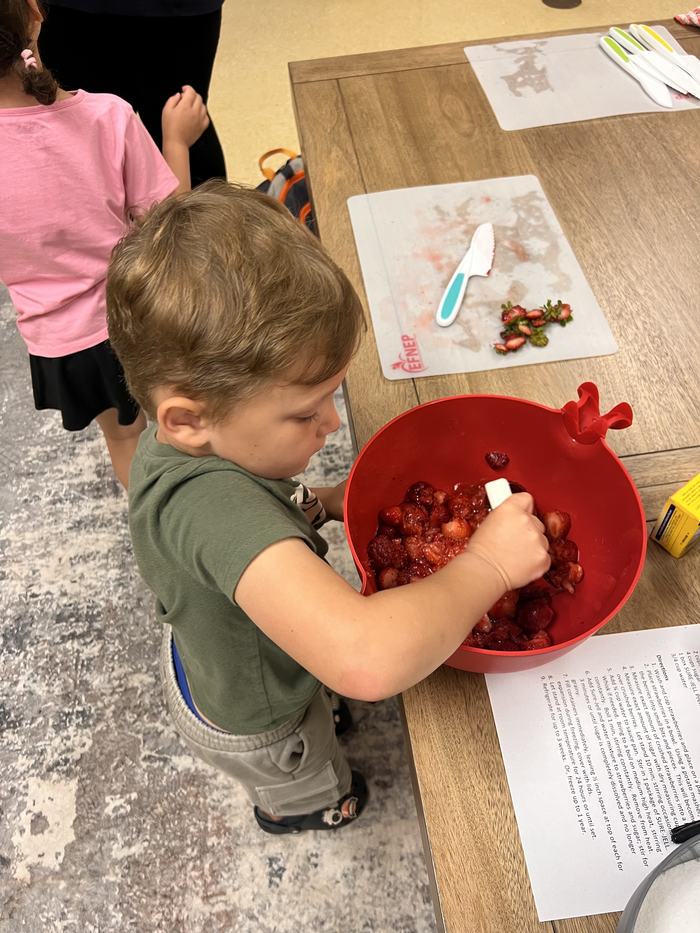 Young child stirring chopped strawberries in a red mixing bowl on a table