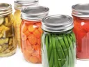 Glass jars of canned vegetables: green beans, sliced carrots, pickles, and tomatoes.