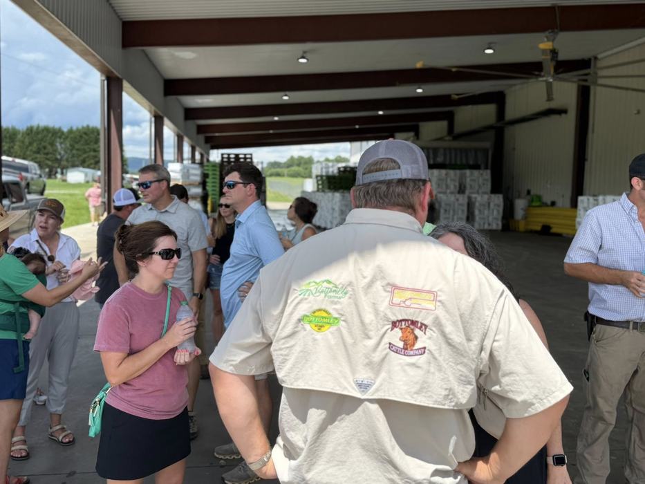 Man in cap wearing logoed shirt talks with group under an open-sided farm shed