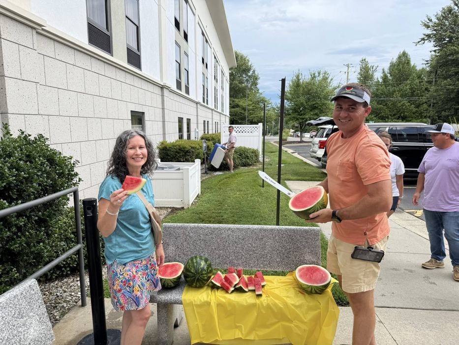 Woman and man holding watermelon slices beside bench with cut watermelons outdoors
