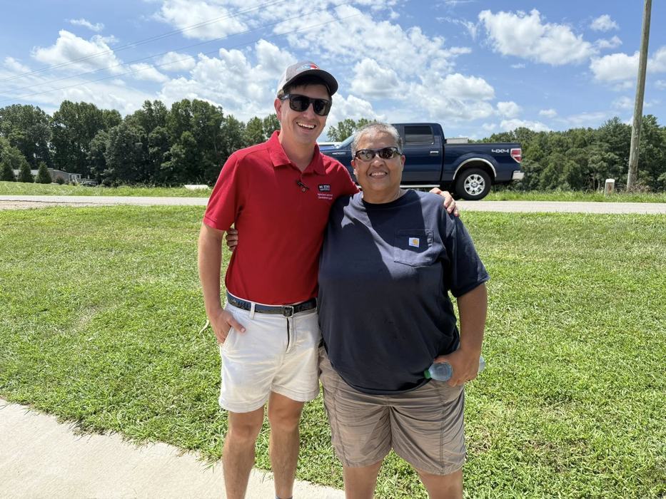 Two people standing arm‑in‑arm on grass; red‑polo and dark‑shirted person holding water bottle; pickup truck behind