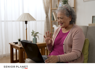 Older woman waving during a laptop video call; SENIOR PLANET from AARP logo
