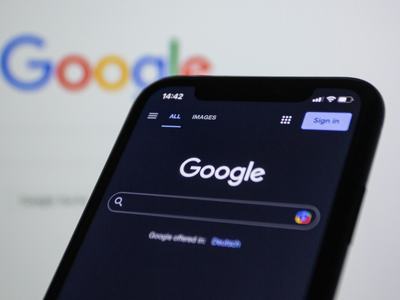 A smartphone showing the Google homepage in dark mode with the search bar and "Sign in" button, set against a blurred background of the Google logo.