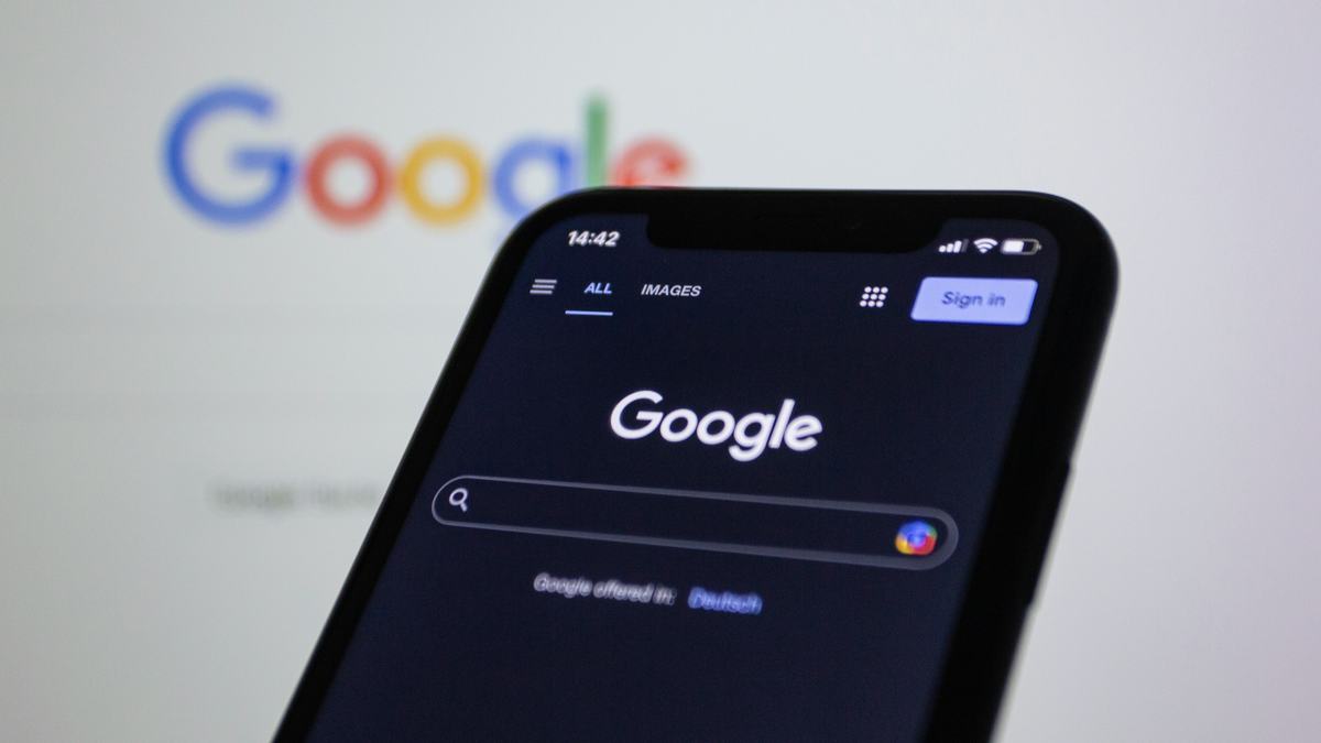 A smartphone showing the Google homepage in dark mode with the search bar and "Sign in" button, set against a blurred background of the Google logo.