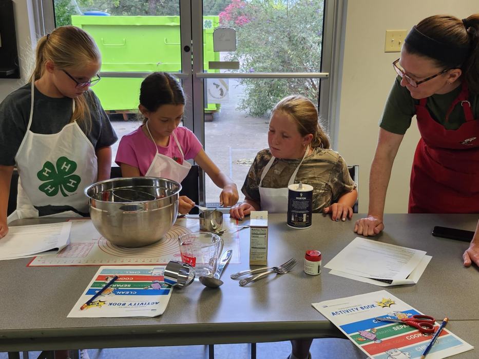 Three girls wearing aprons (one with a 4‑H logo) and a 4-H volunteer gather around a table in a cooking class, measuring ingredients into bowls and consulting activity books and recipe sheets.