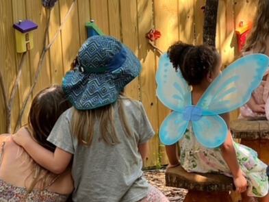 Children wearing colorful wings sit outside and enjoy story time. A young girl is hugging another young girl while they listen.