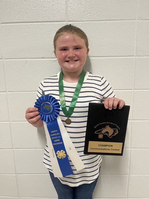 Child holding blue ribbon labeled "North Carolina State 4‑H Horse Program" and plaque "CHAMPION Communications Contest"