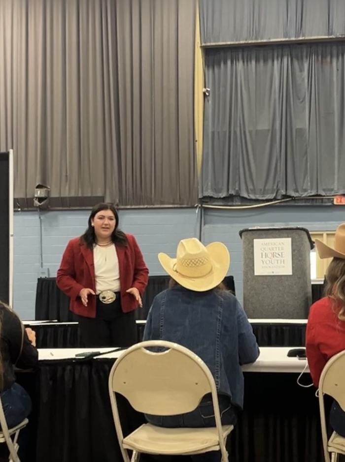 Woman speaking to seated audience; podium sign reads "American Quarter Horse Youth"