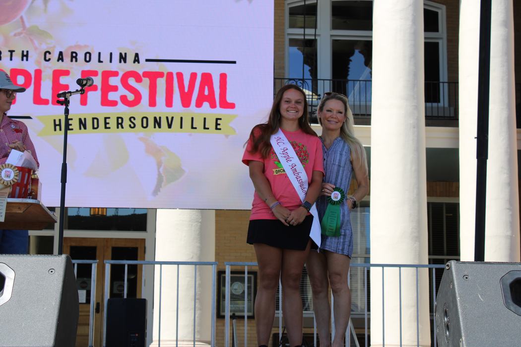 A photo shows two women on an outdoor stage. The woman on the left is wearing a pink T-shirt and a white sash that says "NC Apple Ambassador." The woman on the right is wearing a blue patterned dress and holding a green ribbon. Both are smiling at the camera. In the background, a large screen reads "North Carolina Apple Festival, Hendersonville."