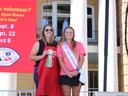 Two women stand on an outdoor stage. The woman on the left wears a red apron and holds a "Grand Champion" ribbon. The woman on the right wears a pink shirt and a sash that says "NC Apple Ambassador." Both are smiling at the camera.