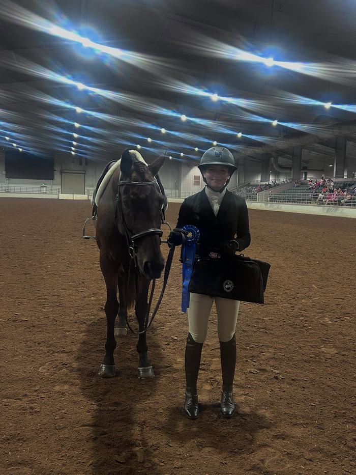 Equestrian rider standing beside horse in indoor arena holding a blue ribbon