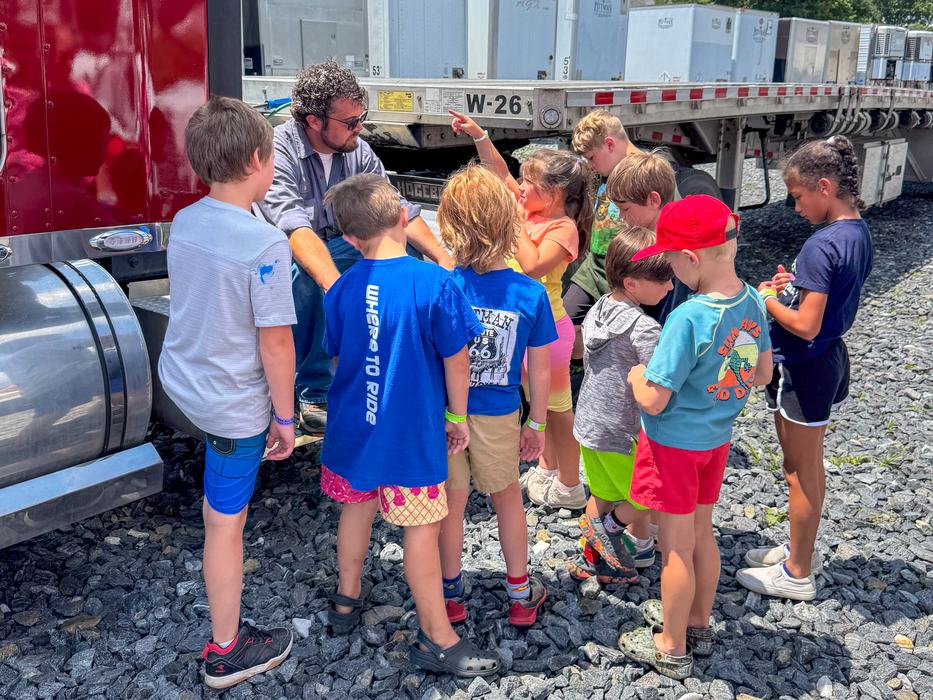 Man kneeling by a truck talking to a group of children gathered on gravel