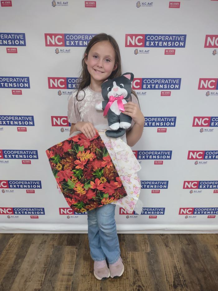 Girl holding stuffed black-and-white cat with pink bow and patterned fabric, NC Extension backdrop.