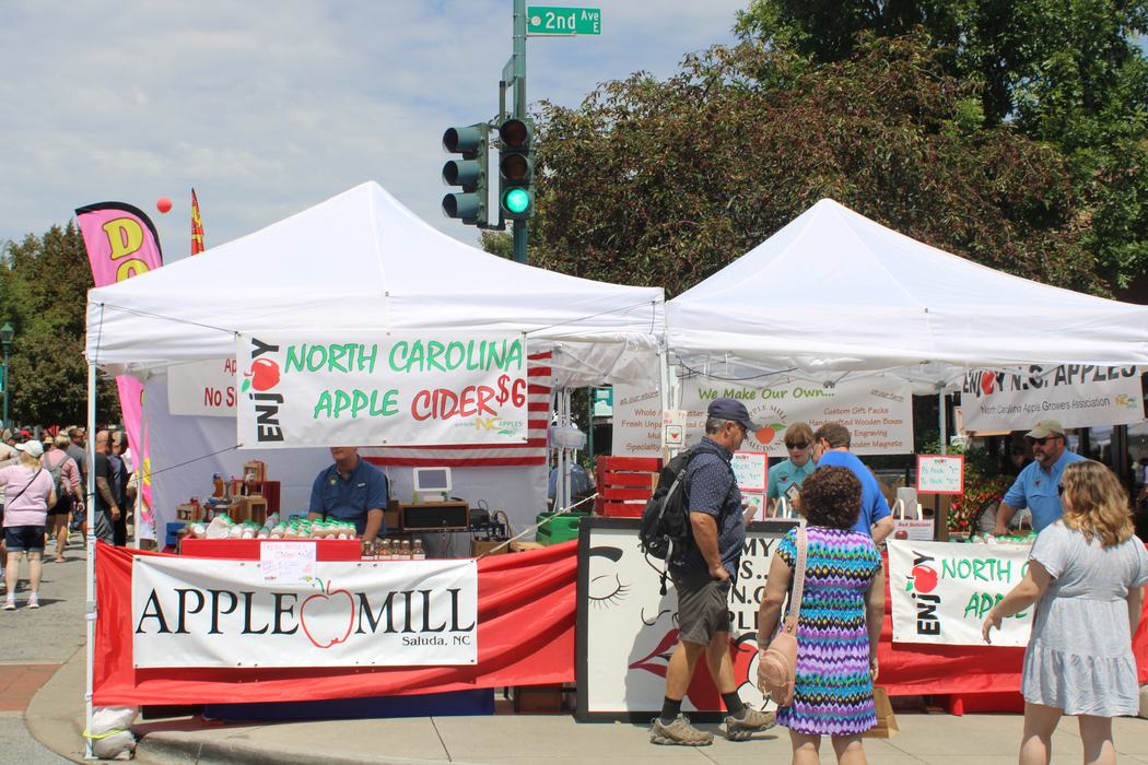 Farmers market tents selling apples and cider; sign "NORTH CAROLINA APPLE CIDER $6"