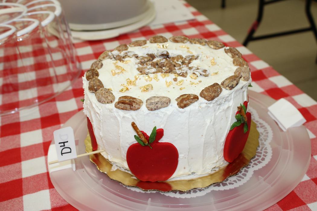 A frosted cake sits on a table with a red and white checkered tablecloth. The cake is decorated with red fondant apples, pecans around the top edge, and what appears to be gold leaf flakes. A small white flag with "D4" written on it is stuck into the side of the cake.