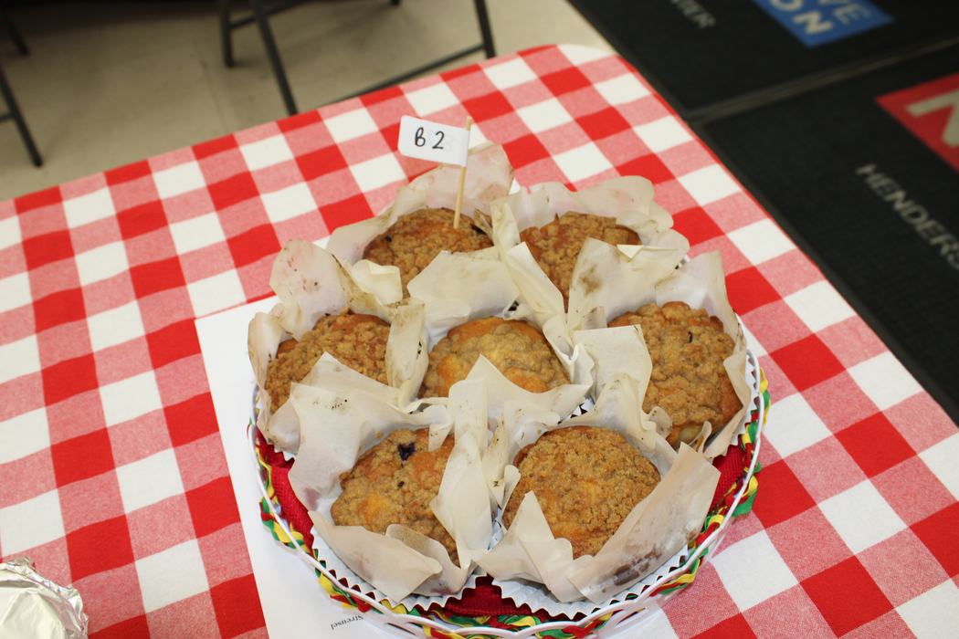 A plate of six muffins in white paper liners sits on a table with a red and white checkered tablecloth. A small white flag with "B2" written on it is stuck in the center of the muffins.
