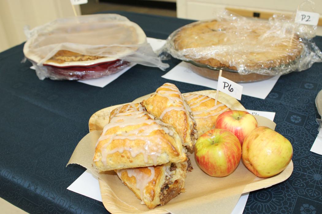  A light-colored plate holds three apple turnovers drizzled with white icing, along with three small apples. A small white flag with "P6" written on it is stuck into one of the turnovers.