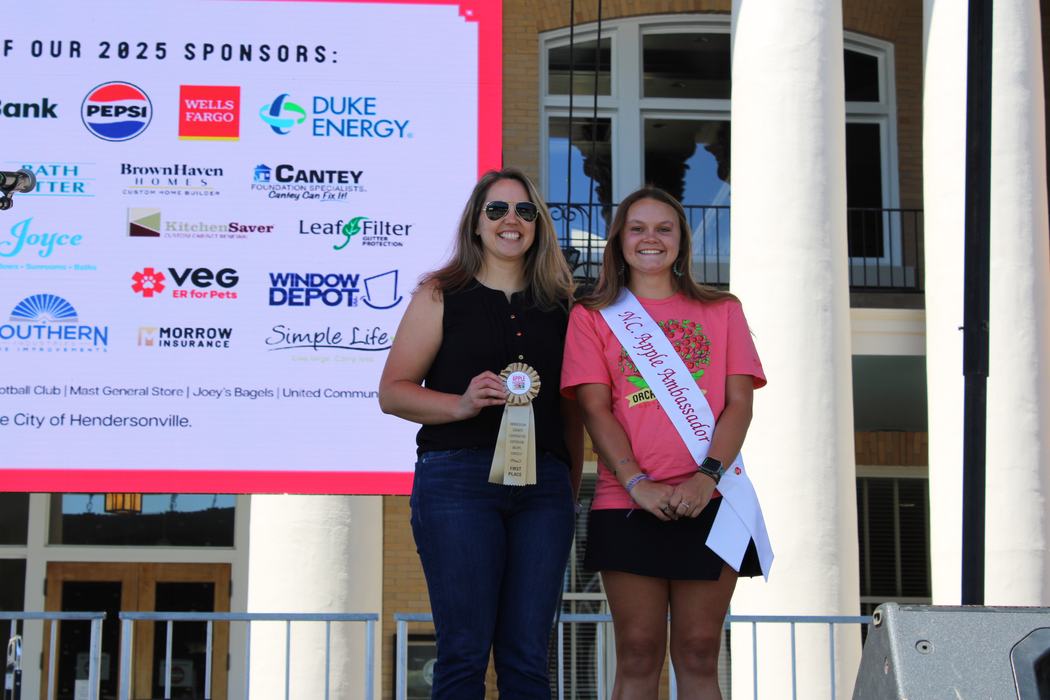 A woman in a black top and a woman in a pink T-shirt stand on a stage, smiling. The woman on the left is holding a ribbon, while the woman on the right is wearing a sash that says "N.C. Apple Ambassador."