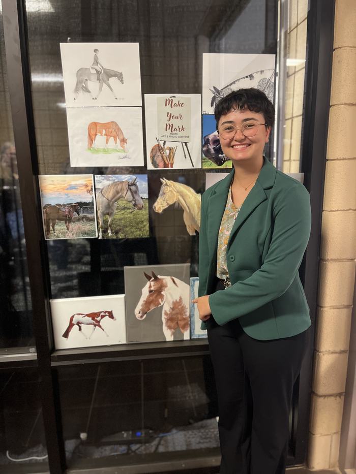 Person in green blazer smiling next to window display of horse artworks and "Make Your Mark" sign.