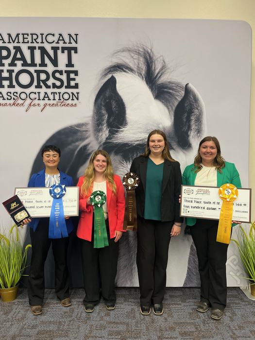 Four young women holding ribbons, checks, and awards in front of "American Paint Horse Association" banner