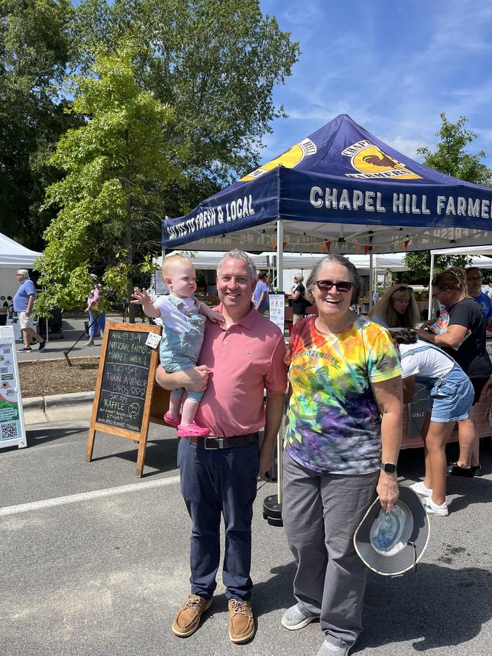 Three people (man holding toddler, woman) standing in front of a tent reading "CHAPEL HILL FARMERS" at a farmers market