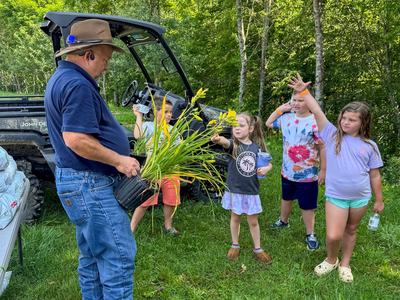 Man holding potted yellow daylilies as four children reach toward them outdoors