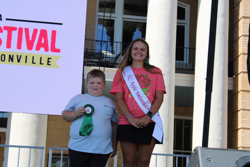 A photo shows a boy and a young woman standing on an outdoor stage. The boy on the left, wearing a light gray T-shirt, holds a green ribbon. The woman on the right, wearing a pink T-shirt and a white "NC Apple Ambassador" sash, smiles at the camera. A large screen behind them reads "Apple Festival."