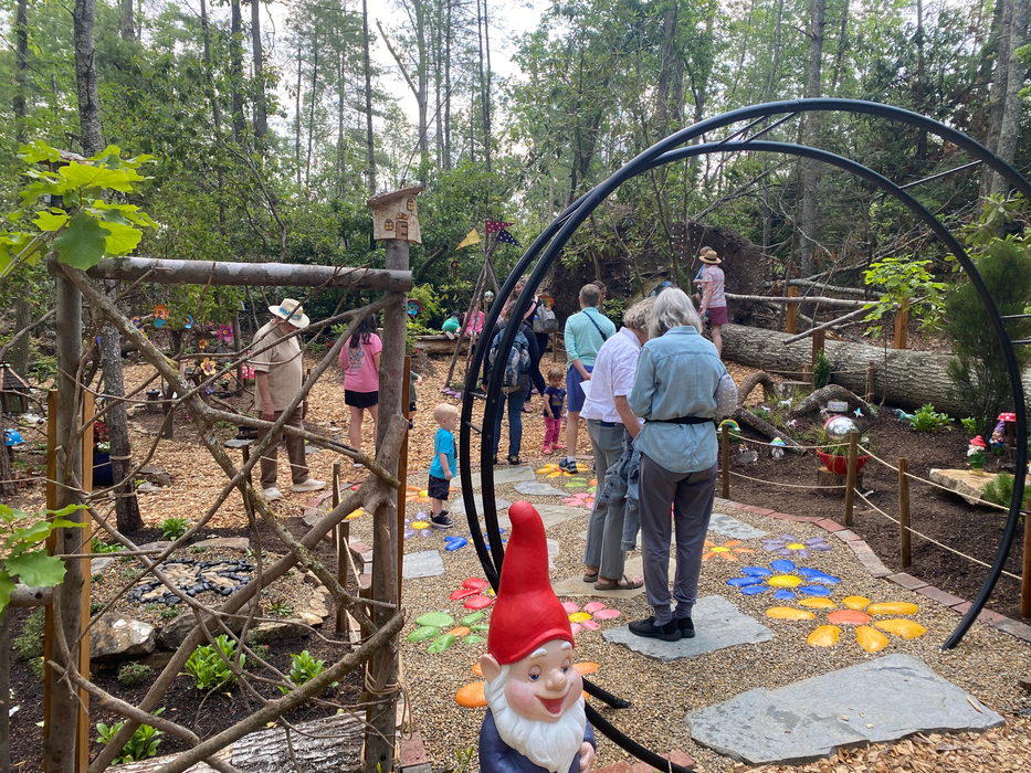 Adults and children walk on an outdoor trail with fairies, gnomes, and flowers.