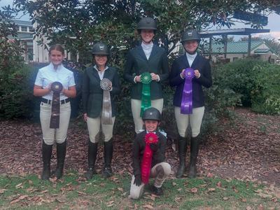 Five equestrians in helmets and show attire holding red, green, purple, silver, and brown ribbons