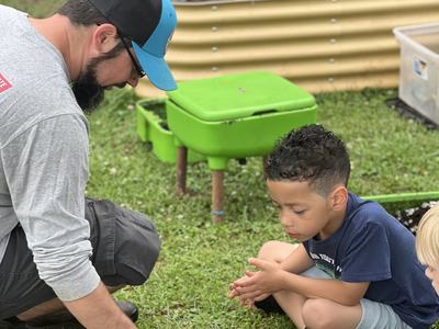Man in blue hat squats down to show a child in a blue shirt the compost in a clear plastic plate. The child sits cross legged looking down at the worms, dirt, and egg shells. In the background are green worm composting bins and a raised bed for gardening.